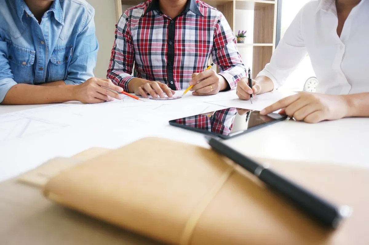 Trois personnes assises autour d'une table comparent des documents et des plans, stylos en main